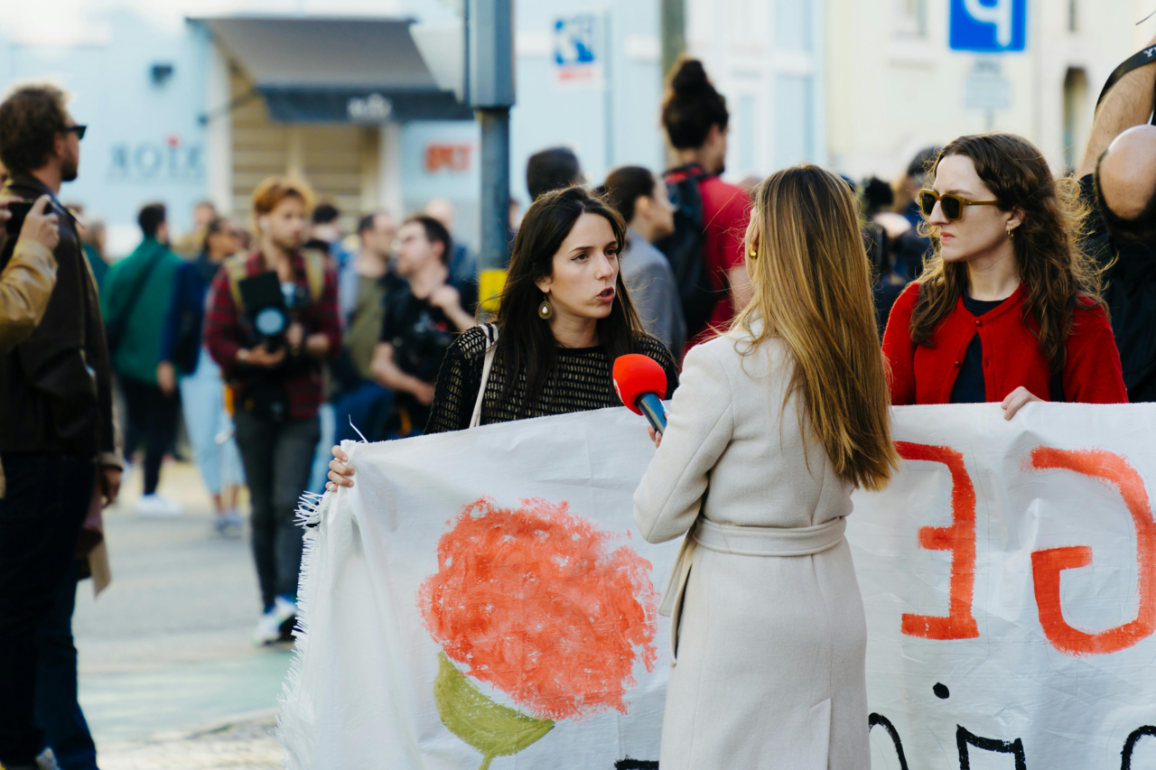 Eine Reporterin in einem beigen Mantel interviewt zwei Frauen, die ein großes Banner halten. Eine der Frauen trägt eine rote Jacke und Sonnenbrille, die andere trägt eine schwarze Bluse und Ohrringe. Im Hintergrund sind weitere Personen zu sehen, die sich in einer belebten städtischen Umgebung versammelt haben.